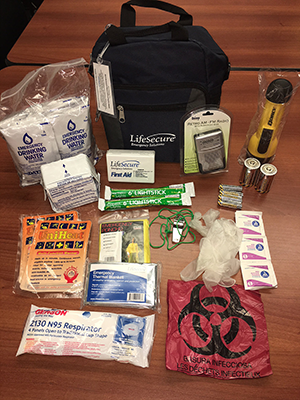 An image of an emergency kit with preparedness items laid out on a table. Items consist of a flashlight, emergency water, emergency food, a radio, a first aid kit, glow sticks, hand warmers, batteries, a biohazard bag, medical gloves, an N95 respirator, a mylar blanket, alcohol prep wipes, a poncho, and a metal whistle.